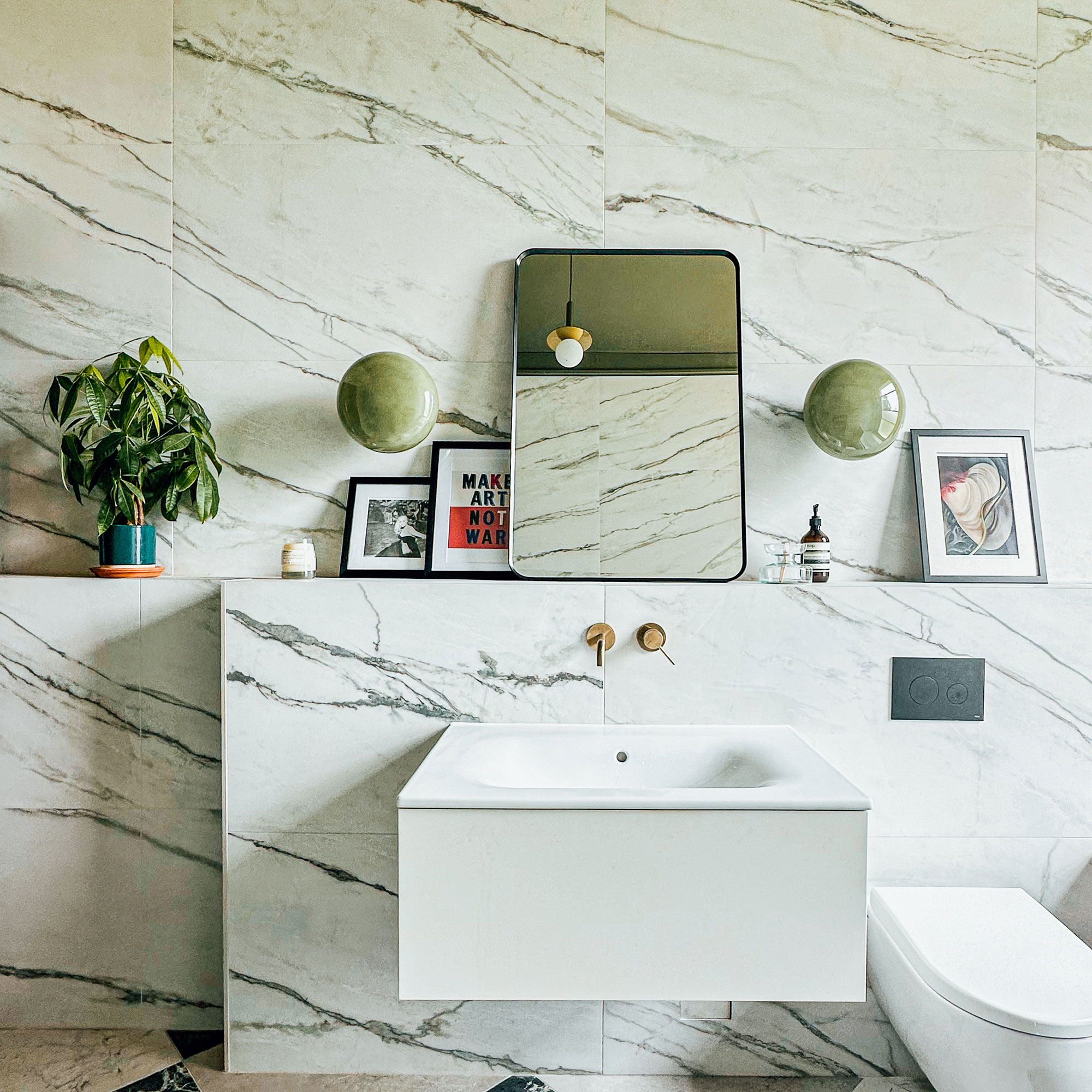 Bathroom with marble walls, floating sink, open shelf above with artwork, plant and mirror and two round green wall lights