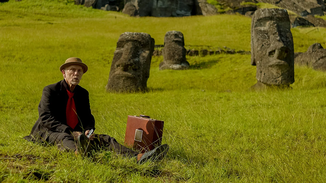 An elderly man (John Malkovich as Chris) sits in the grass next to Easter Island heads, in 'Wild Horse Nine.'