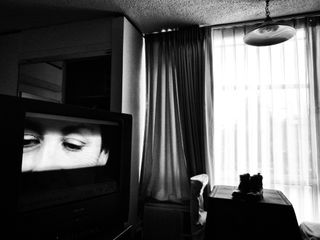 Black and white photograph of a room with a large TV showing a close up of a person’s eyes.