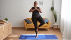 woman in black vest and leggings standing in tree pose on a blue yoga mat in a living room. there's a yellow sofa behind her
