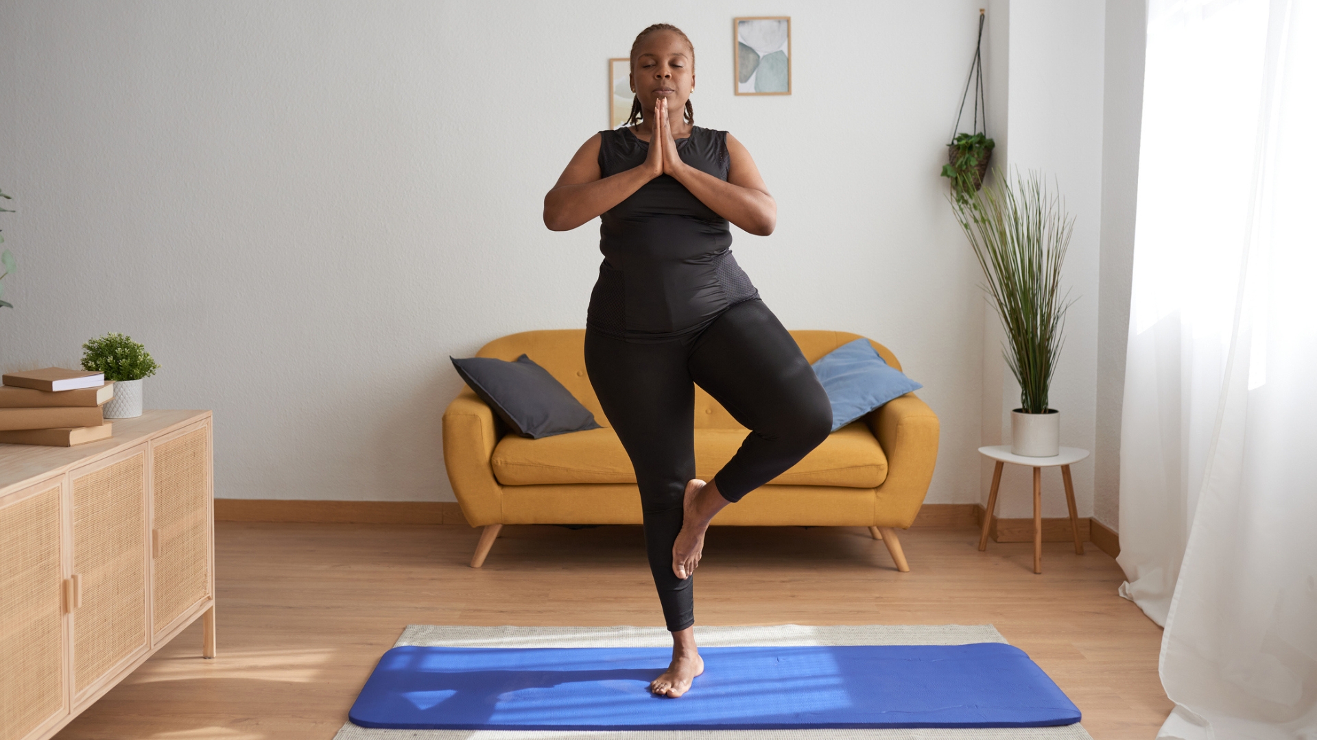 woman in black vest and leggings facing the camera standing in tree pose on a blue yoga mat in a living room. there&#039;s a yellow sofa behind her