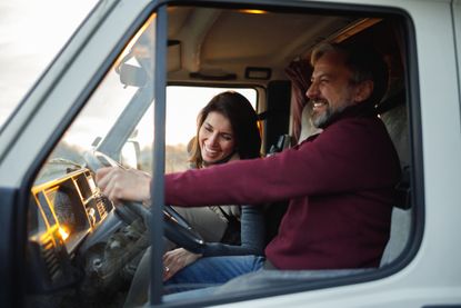 A mid-age couple is traveling with the RV. The husband is driving, and the wife sits next to him. They are laughing.