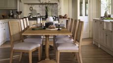 A light wood kitchen table in the centre of a white kitchen with six chairs