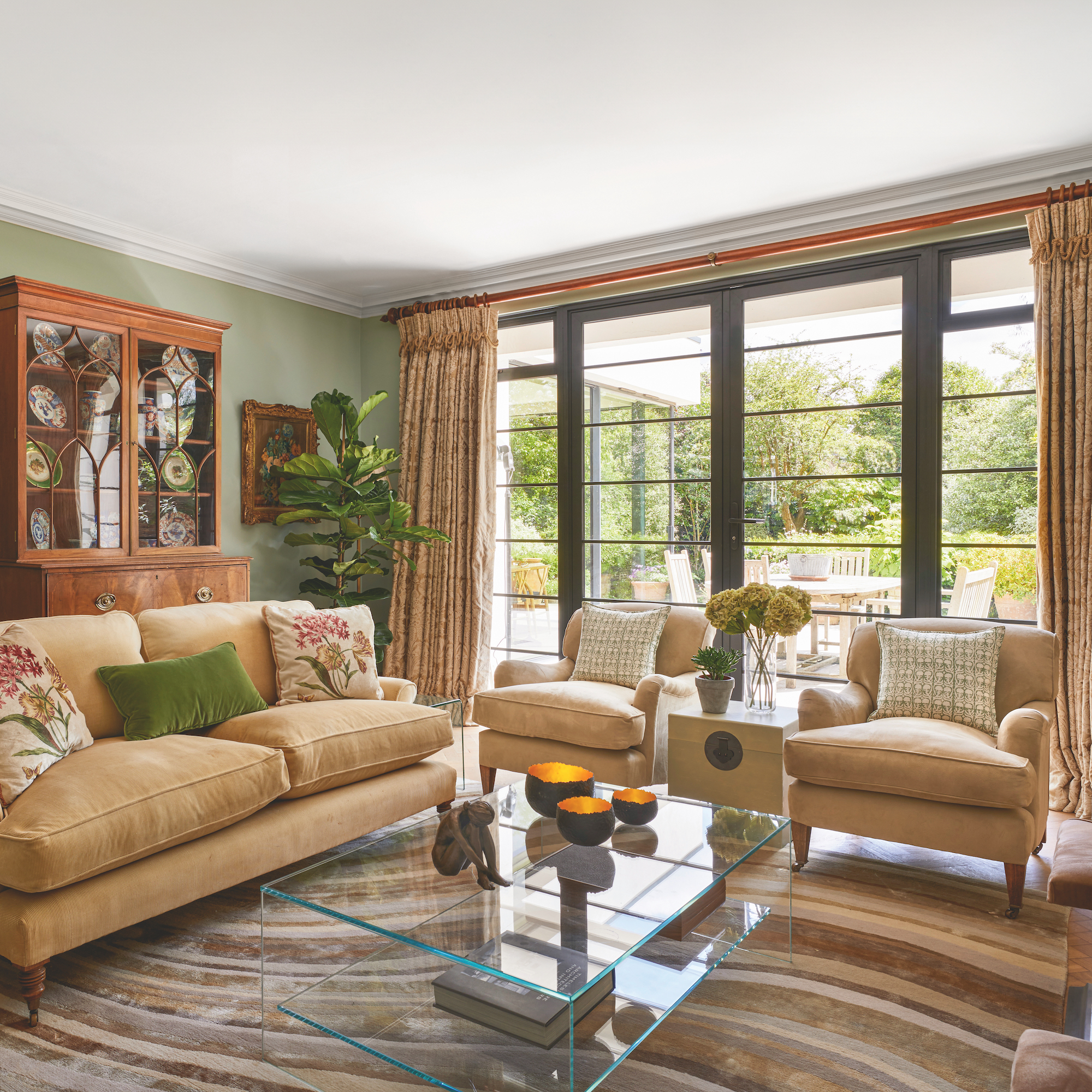 Sitting room with Crittall windows and pale gold armchairs and sofa surrounding glass coffee table