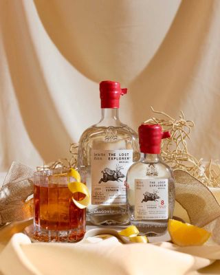 Two bottle of mezcal with a red, glazed top stand to the right of a filled glass covered in lemon peel swirls against a beige background.