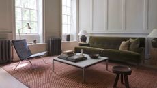 Image of a white living room with a brick red jute rug, an olive green sofa, a black leather accent chair, and a square coffee table.