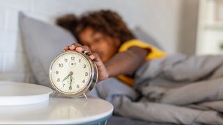 A woman with afro hair reaching out of bed to turn her alarm clock off on light mornings as clocks go back.