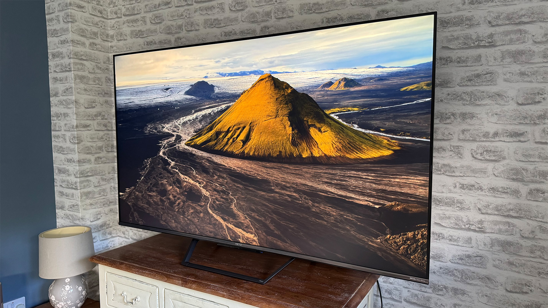Metz MQE7600 4K TV on wooden sideboard in front of brick wall, on screen is mountainous landscape
