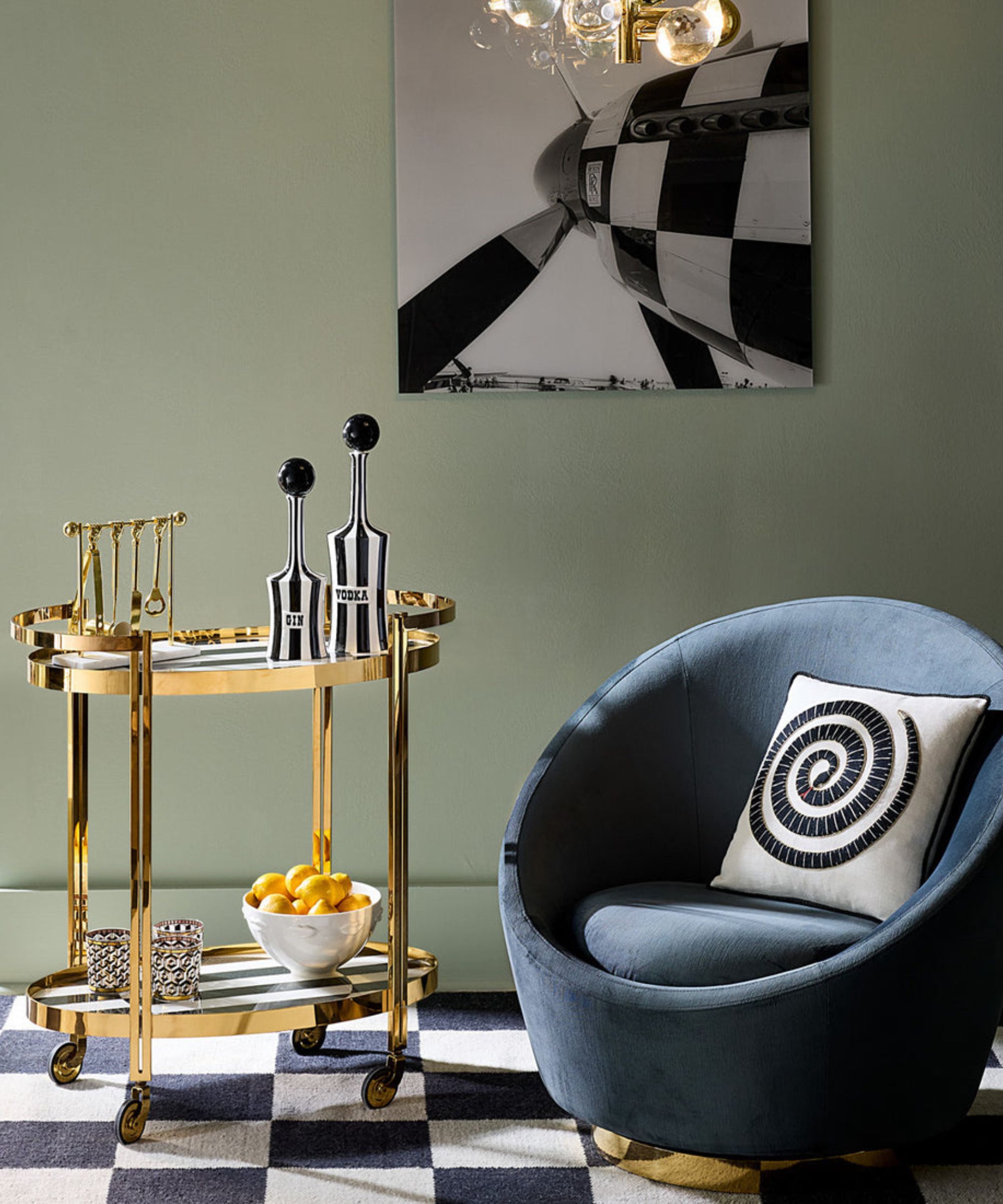 A chic lounge corner featuring a gold bar cart with "Gin" and "Vodka" decanters, a blue velvet swivel chair, and a black-and-white checkered rug