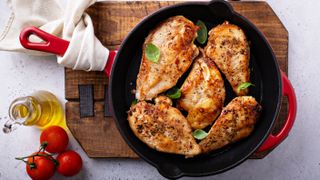 A red enameled cast iron skillet which contains five chicken breasts on a wooden platter