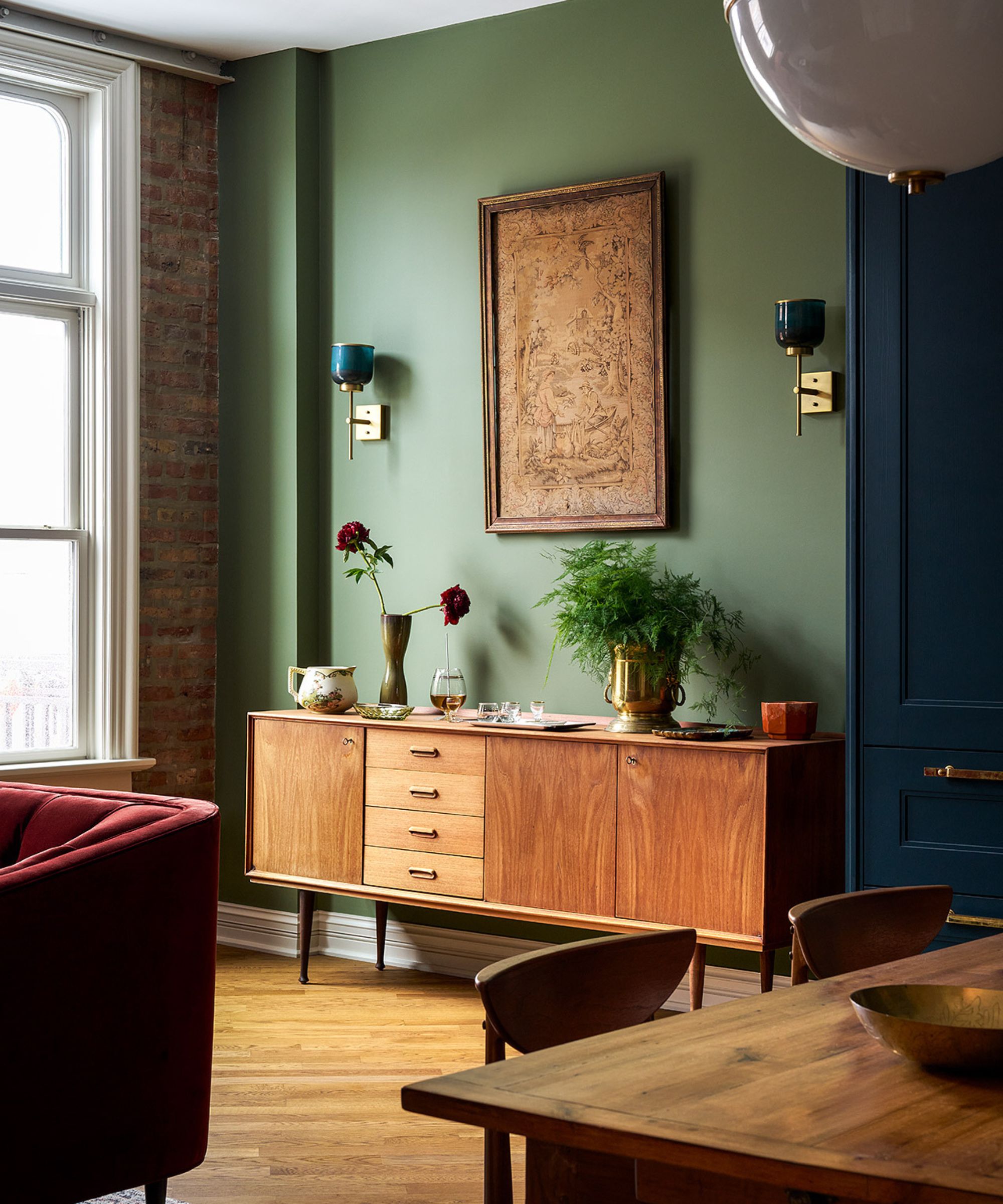 A modern living room with a mossy green wall, wooden flooring, a wooden sideboard with artwork above it, and a brick wall with a window.