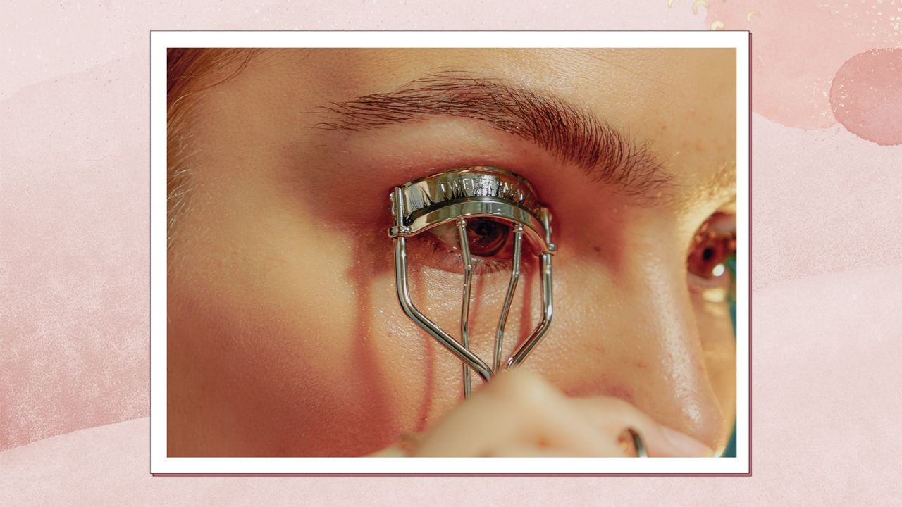 A close up of a woman&#039;s eye and eyebrow as she curls her eyelashes with an eyelash curler/ in a pink textured watercolour-style template