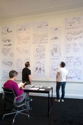 Two men looking at sketches on wall while one man sits at desk