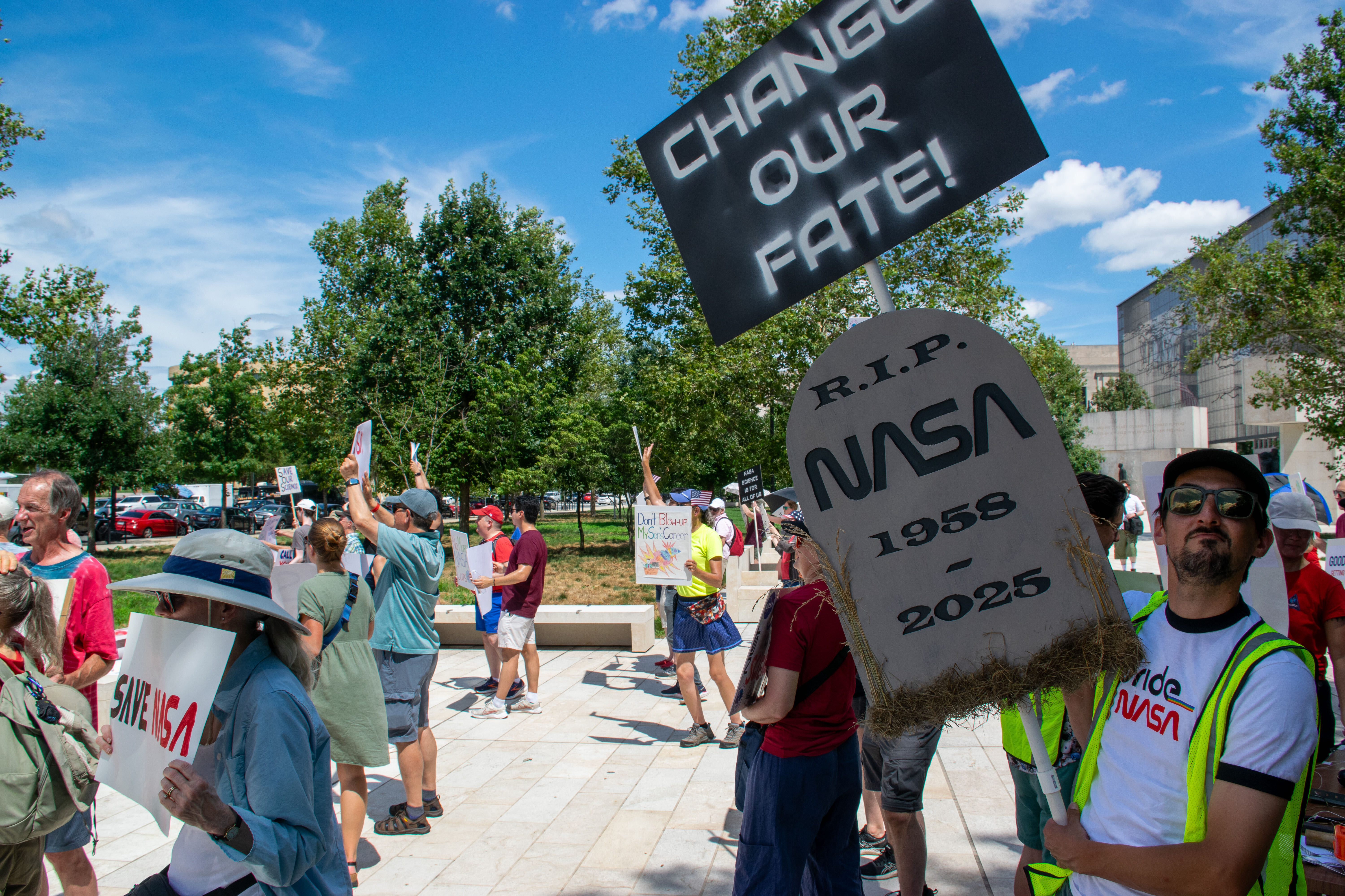 People hold signs outside on a sunny day in protest of NASA budget cuts, July 20, 2025.