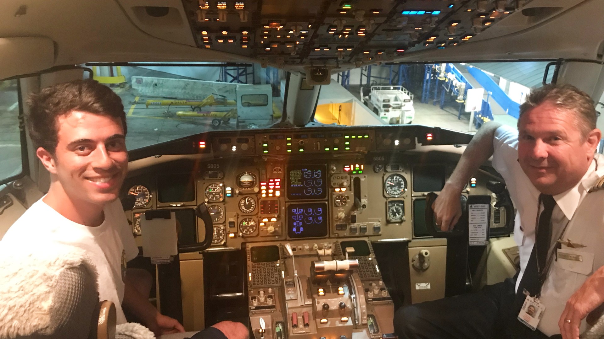 Nick is shown in the cockpit of a plane with the pilot. They are both smiling and looking towards the camera.