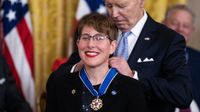 a woman wearing a nasa pin smiles with red lipstick as an old man in a suit behind her pins the neckalce of a medal hanging around her neck.