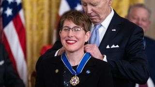 a woman wearing a nasa pin smiles with red lipstick as an old man in a suit behind her pins the neckalce of a medal hanging around her neck.