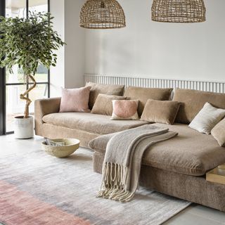 living room with large stone L-shaped sofa, pair of rattan pendants, rug, pink and stone cushions, plant, white walls, crittall doors