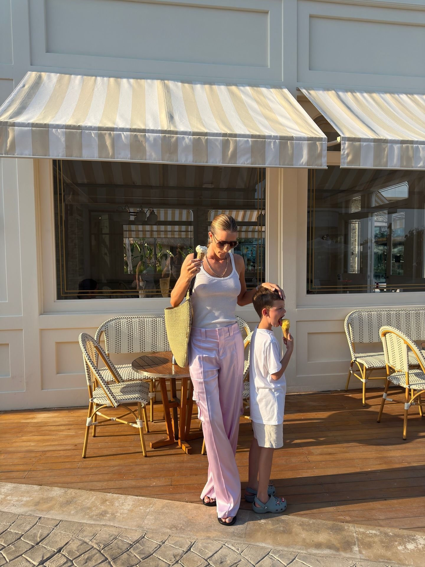 Fashion person Rosie Huntington-Whiteley wears the pale pink trouser trend. She pairs her trousers with a white tank top, khaki raffia tote and thong sandals on holiday. She is looking down, holding her son's head in front of a old-fashioned ice-cream shop in a recent image from her Instagram.