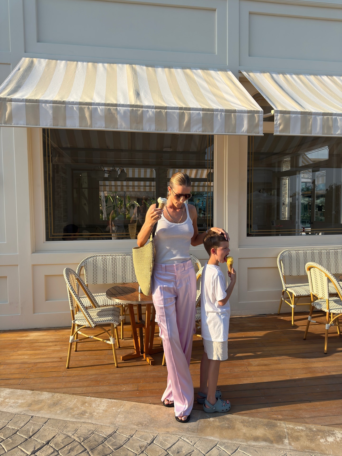 Fashion person Rosie Huntington-Whiteley wears the pale pink trouser trend. She pairs her trousers with a white tank top, khaki raffia tote and thong sandals on holiday. She is looking down, holding her son's head in front of a old-fashioned ice-cream shop in a recent image from her Instagram.