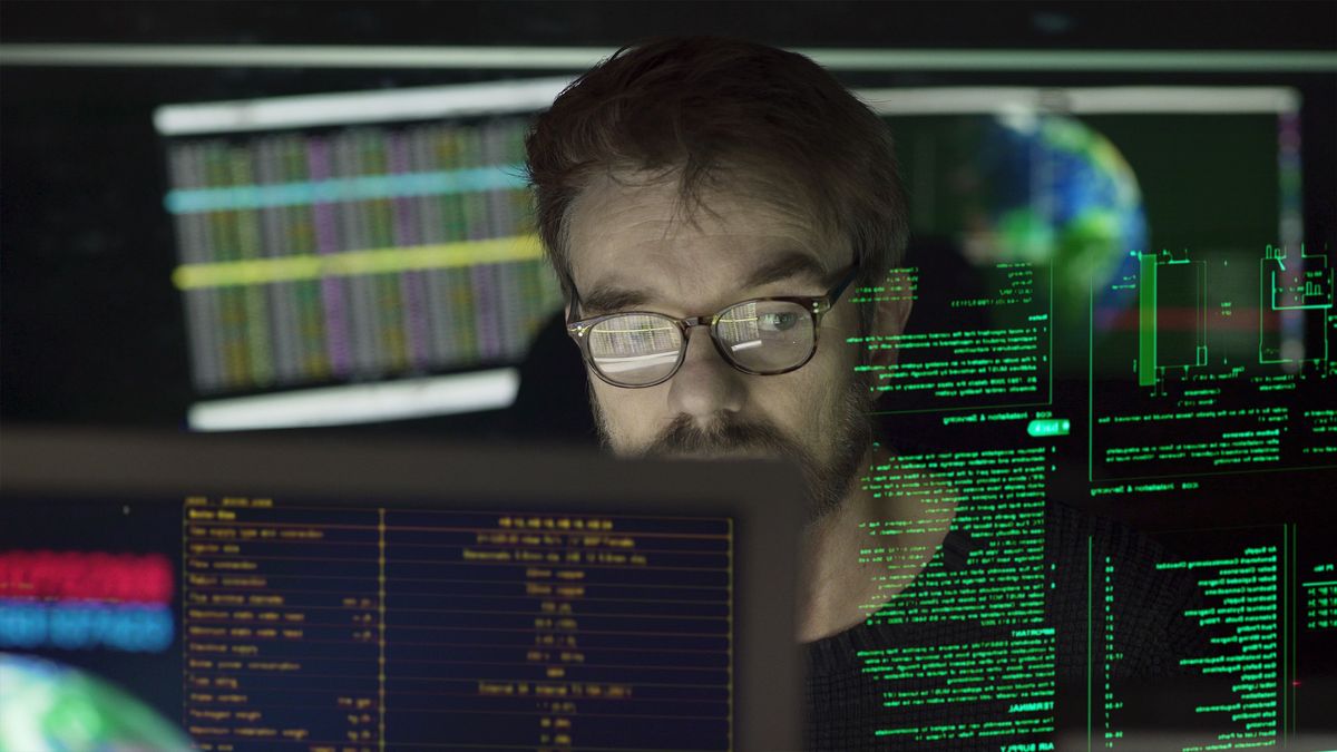 A man surrounded by monitors and holographic displays showing lines of code