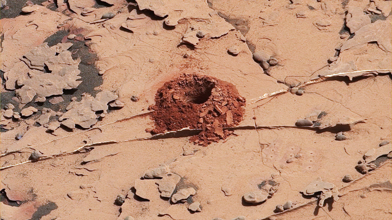 Rusty red Martian landscape, close up of a small hole drilled by Curiosity with a mound of rusty red material surrounding it.