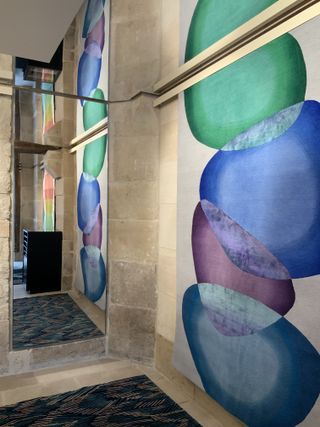 Interior view of Deirdre Dyson’s showroom featuring a long blue, green, purple and grey rug hanging from a stone wall in front of a mirror. The mirror shows the reflection of that rug along with a black unit and another long multicoloured rug hanging on the wall. There is a third dark coloured patterned rug on the floor which can also be seen in the mirror