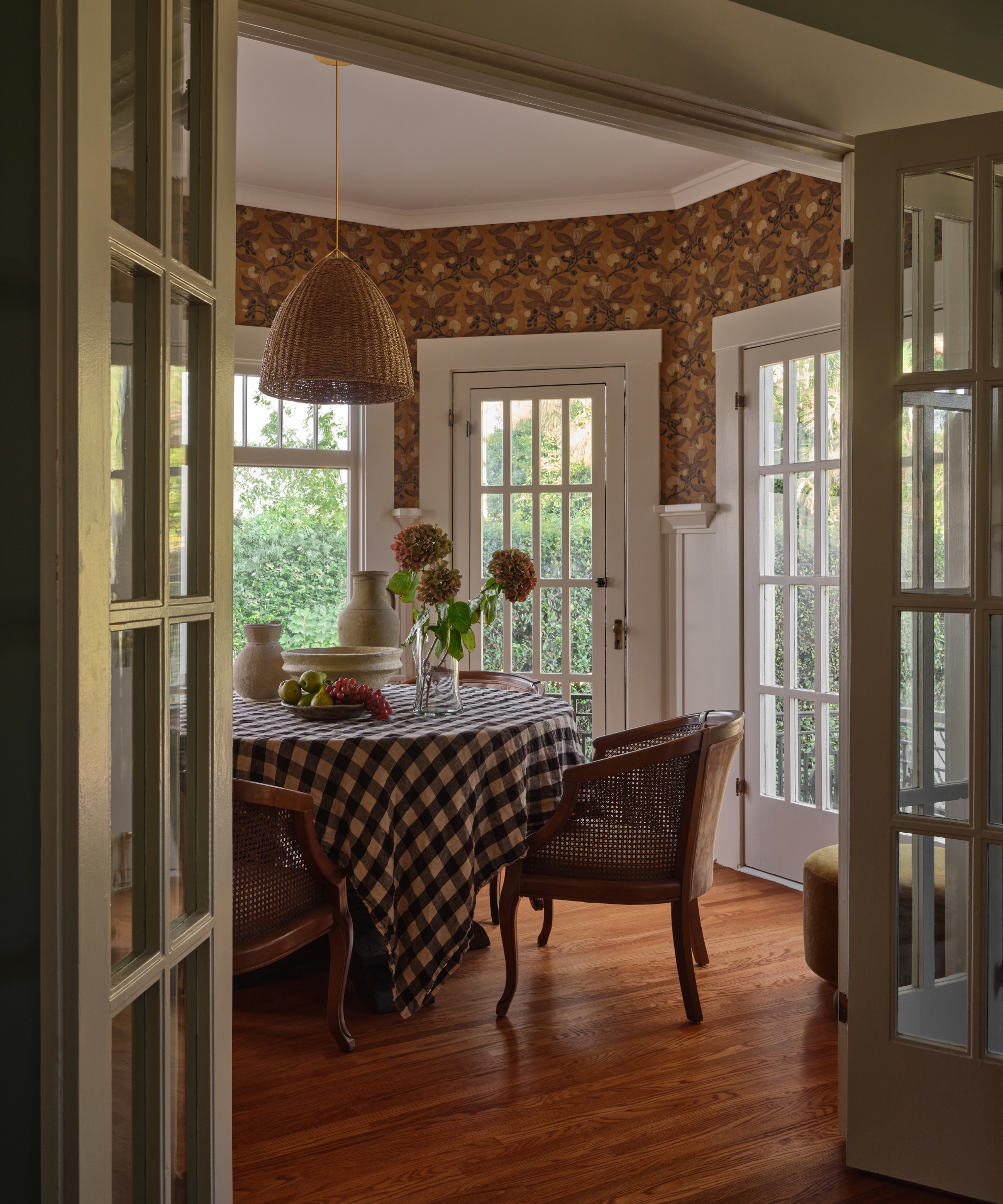 A breakfast room connected to a galley kitchen, featuring wallpapered walls, a round table covered in a blue and white gingham tablecloth, and can chairs