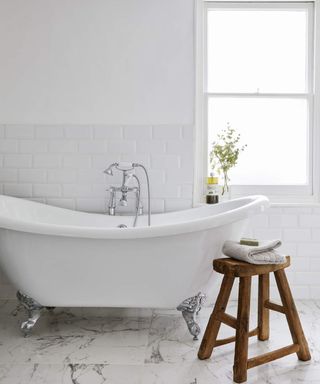 A white bathroom with small white bathtub, marble flooring and small wooden stool