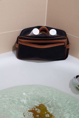 Black makeup bag on the edge of a white bathtub filled with water.