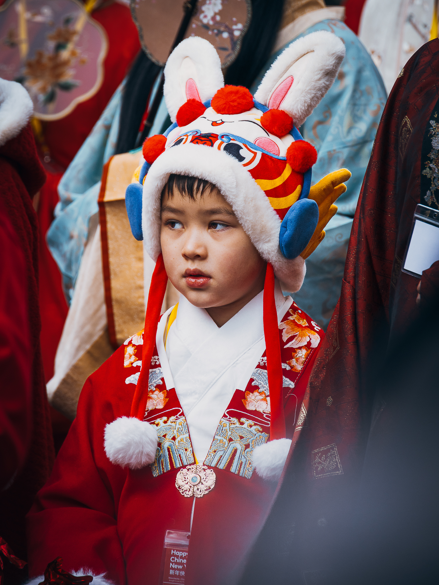 A young child in traditional attire wears a colorful rabbit hat with fluffy ears and red pom-poms, surrounded by people in festive clothing