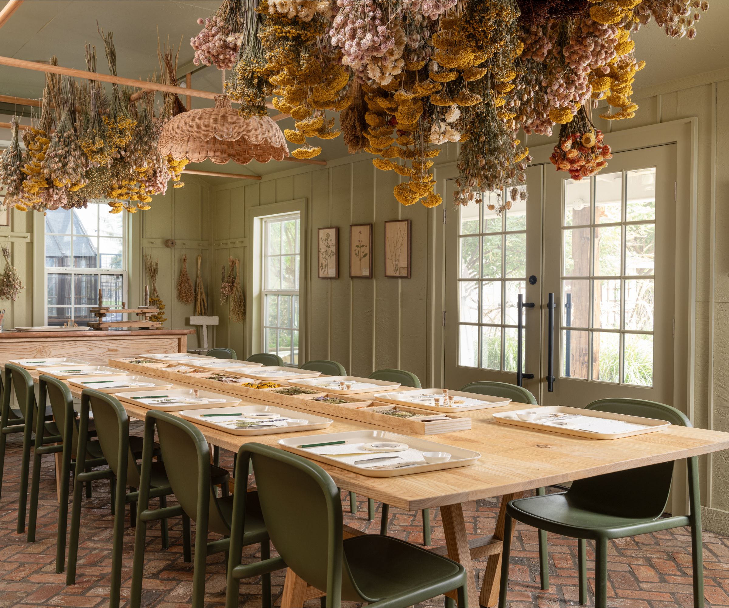 Room with brick floor, sage green painted walls, dried flowers hanging on ceiling, rattan light and long wood dining table with green chairs