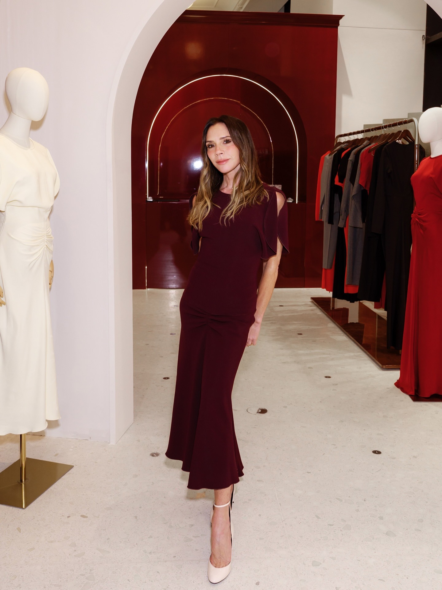 Victoria Beckham stands inside her shop wearing a burgundy dress with cream heels.