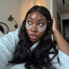 A woman taking a selfie in a hotel robe wearing under-eye patches with dark brown, glossy hair