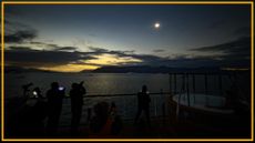 A view of people standing on a beach looking up at a dim sun with a yellow border around the image.