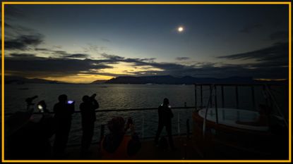 A view of people standing on a beach looking up at a dim sun with a yellow border around the image.