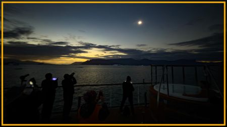 A view of people standing on a beach looking up at a dim sun with a yellow border around the image.