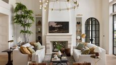 A bright living room with two white sofas facing a stone fireplace. A large tree sits in the corner, and shelves with pottery line the fireplace. A round gold chandelier hangs above, and tall black-framed windows let in natural light. A coffee table with books and flowers sits in the center.