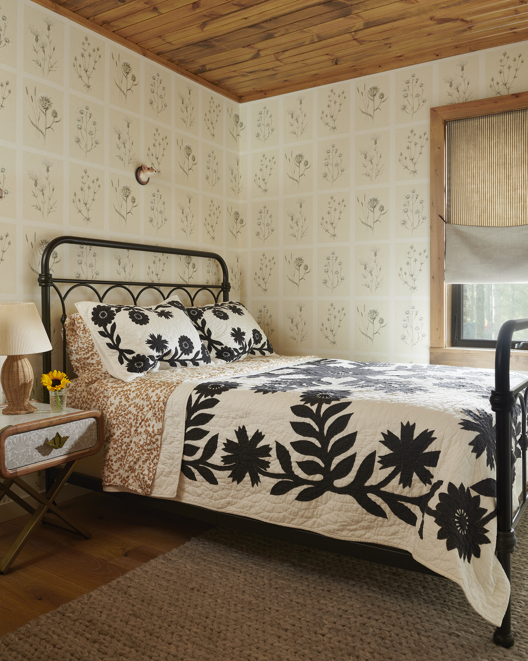 A bedroom with a black metal bed that has a black and white patchwork quilt on it. The walls are covered in a patchwork-inspired wallpaper and there is a brown woven rug on the floor.