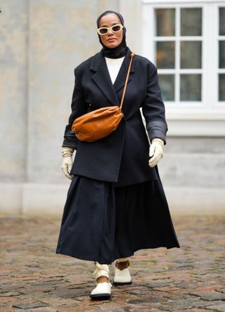 A guest wears a white sunglasses, a black scarf as a headband, a white t-shirt, a black buttoned coat, a camel shiny leather crossbody bag, white latte leather gloves, white latte leather ballerinas , outside Marimekko, during the Copenhagen Fashion Week Spring/Summer 2024 on August 09, 2023 in Copenhagen, Denmark.