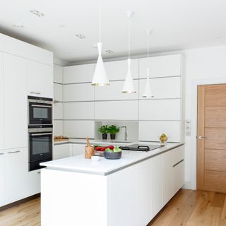 Kitchen with white cabinets, peninsula, and hanging lamps, wood floor and door, black induction hob and twin ovens, and colourful decorative touches