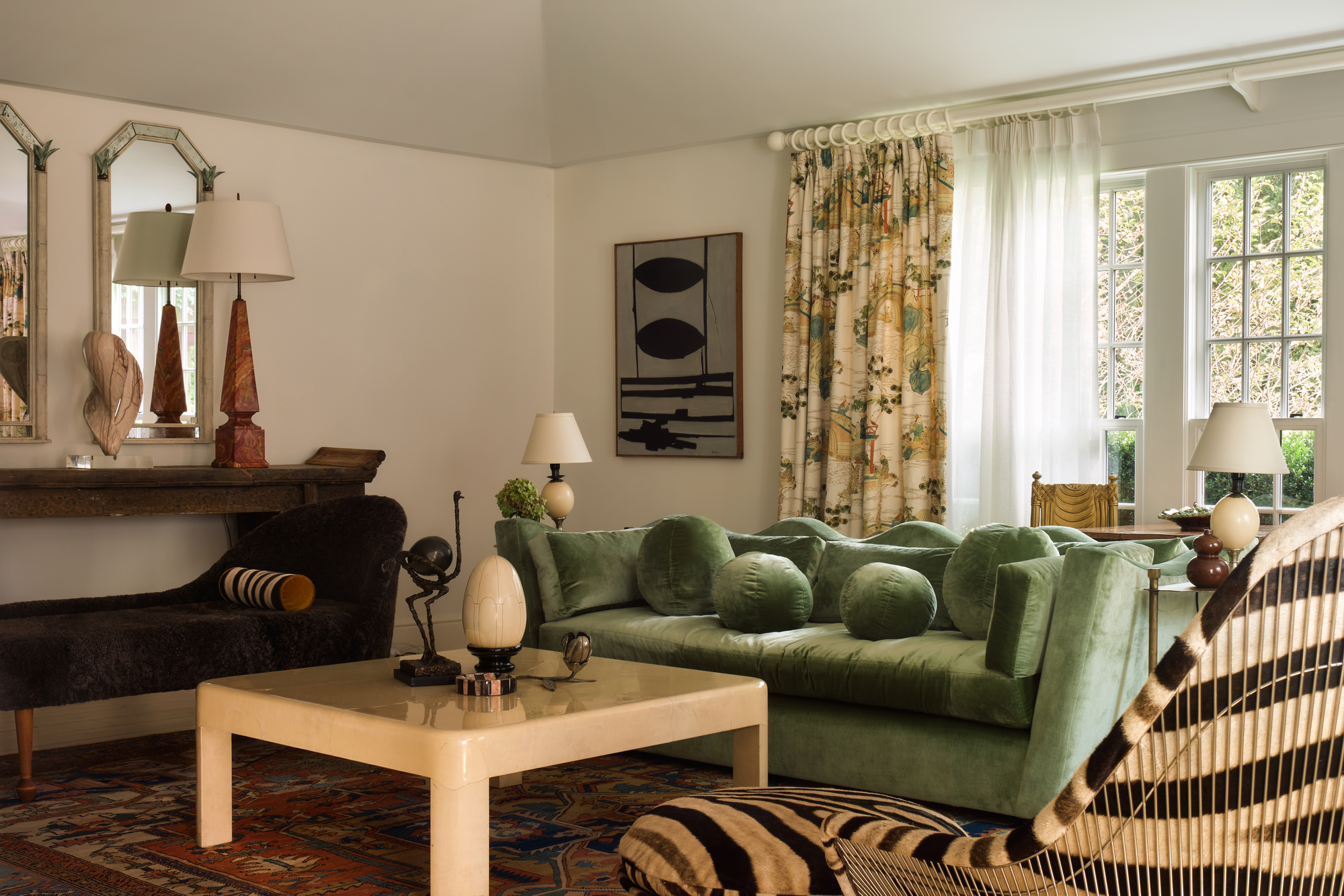 modern living room with green velvet sofa and wavy backrest, floral curtains layered over sheets, a console table styled with two mirrors, a lamp, and a bust, a square coffee table styled with small decor and a patterned rug on the floor