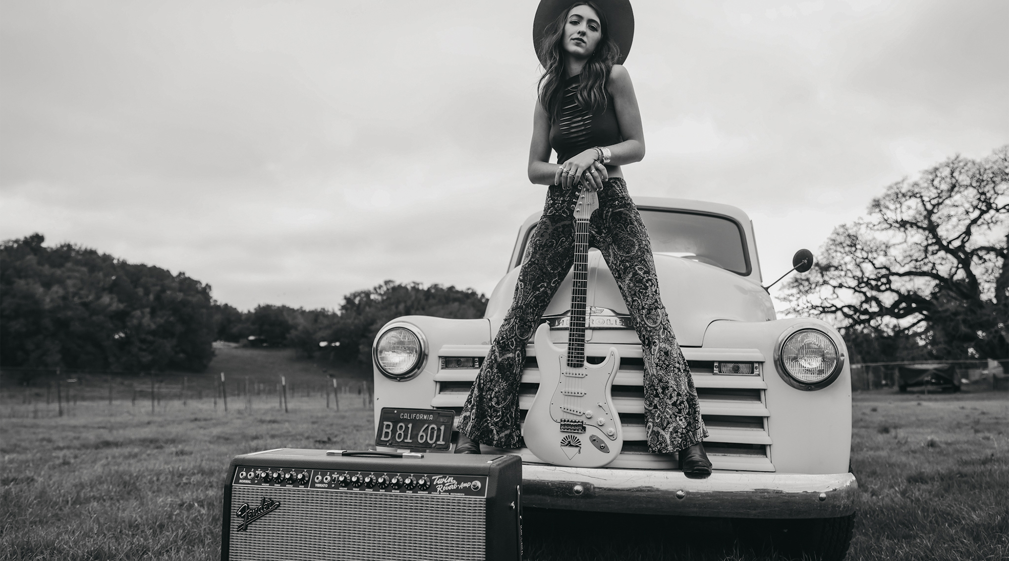 A photo of guitarist Bella Rayne standing on the bumper of a vintage Chevrolet auto with her Fender Stratocaster. A Fender Twin Reverb amplifier is in the foreground.