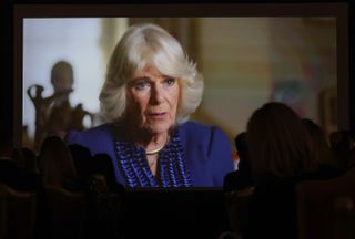 An image of Queen Camilla wearing a blue dress on a large screen as silhouettes of viewers watch her documentary in an audience