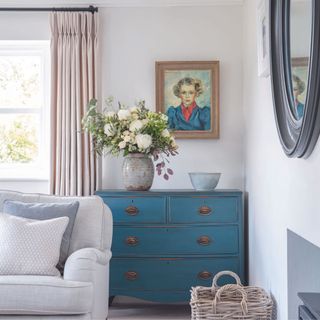 corner of living room with grey walls and sofa and dark blue chest of drawers with flowers and painting of a woman above
