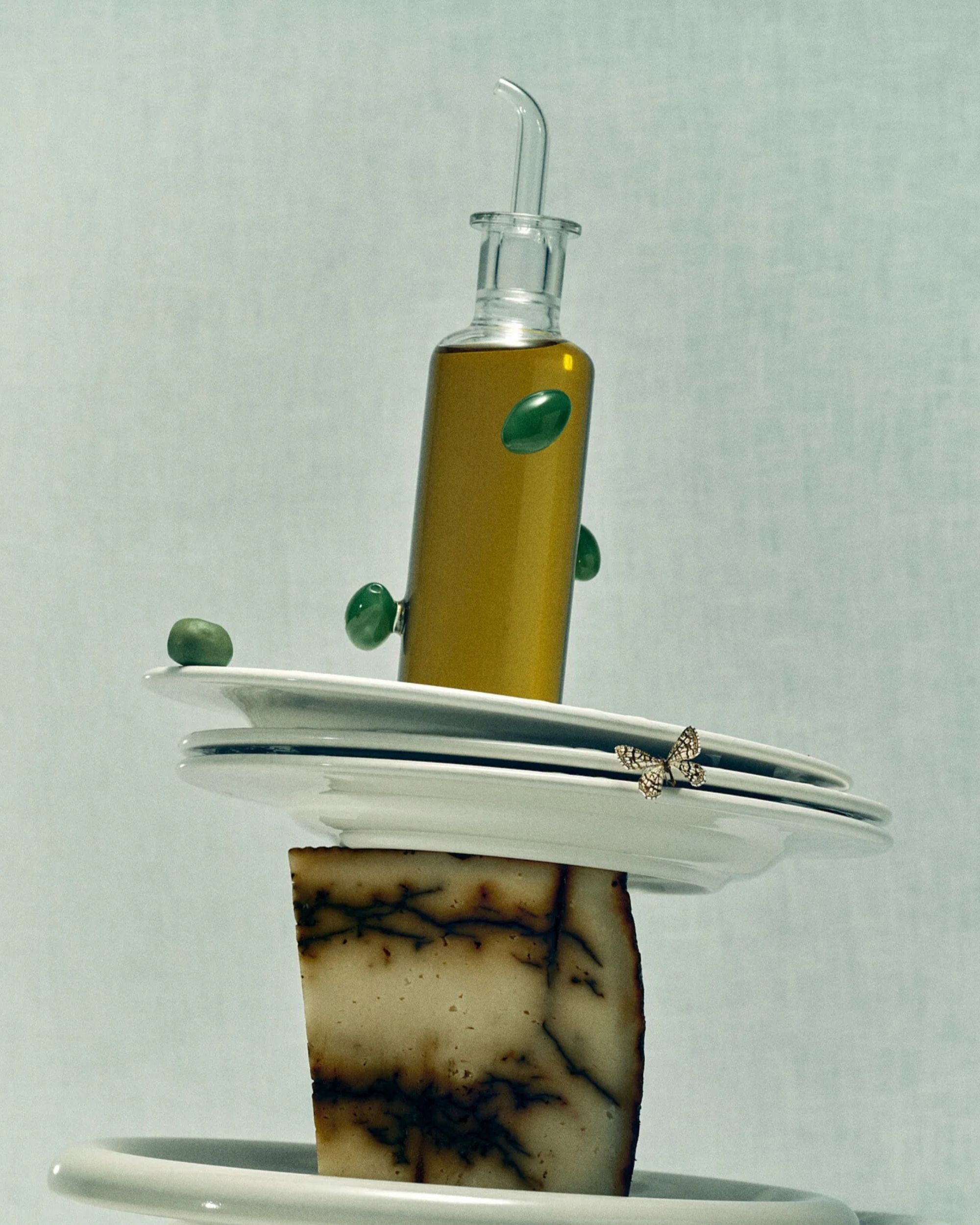 Image of a glass olive oil holder with olives on it, balancing on a stack of white plates being held up by cheese.