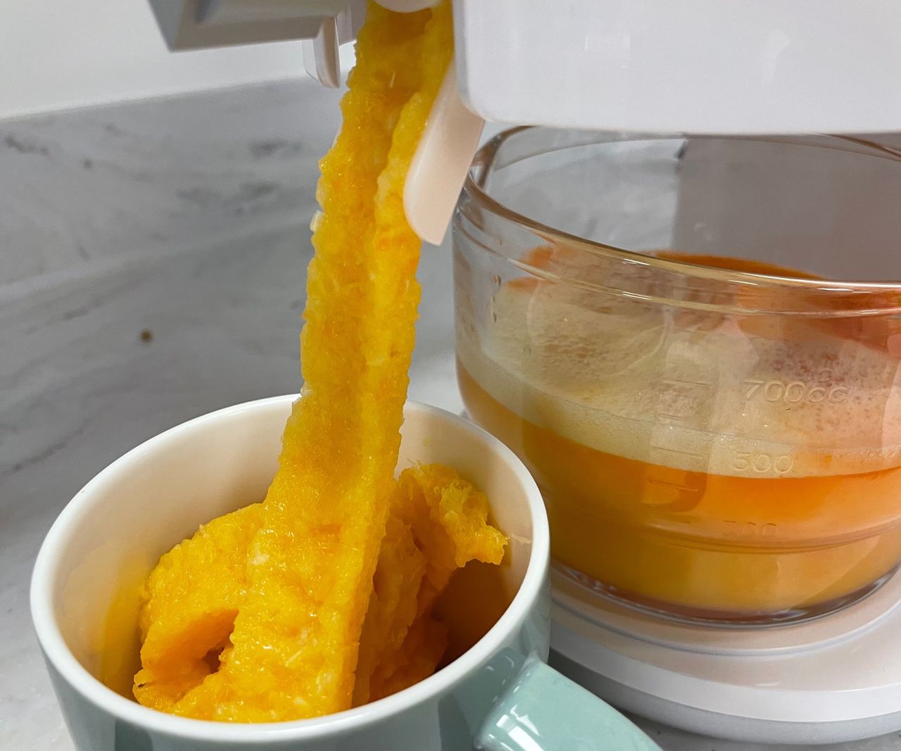 Orange fruit pulp being pushed from a white juicer into a ceramic mug, beside a glass beaker of orange liquid.