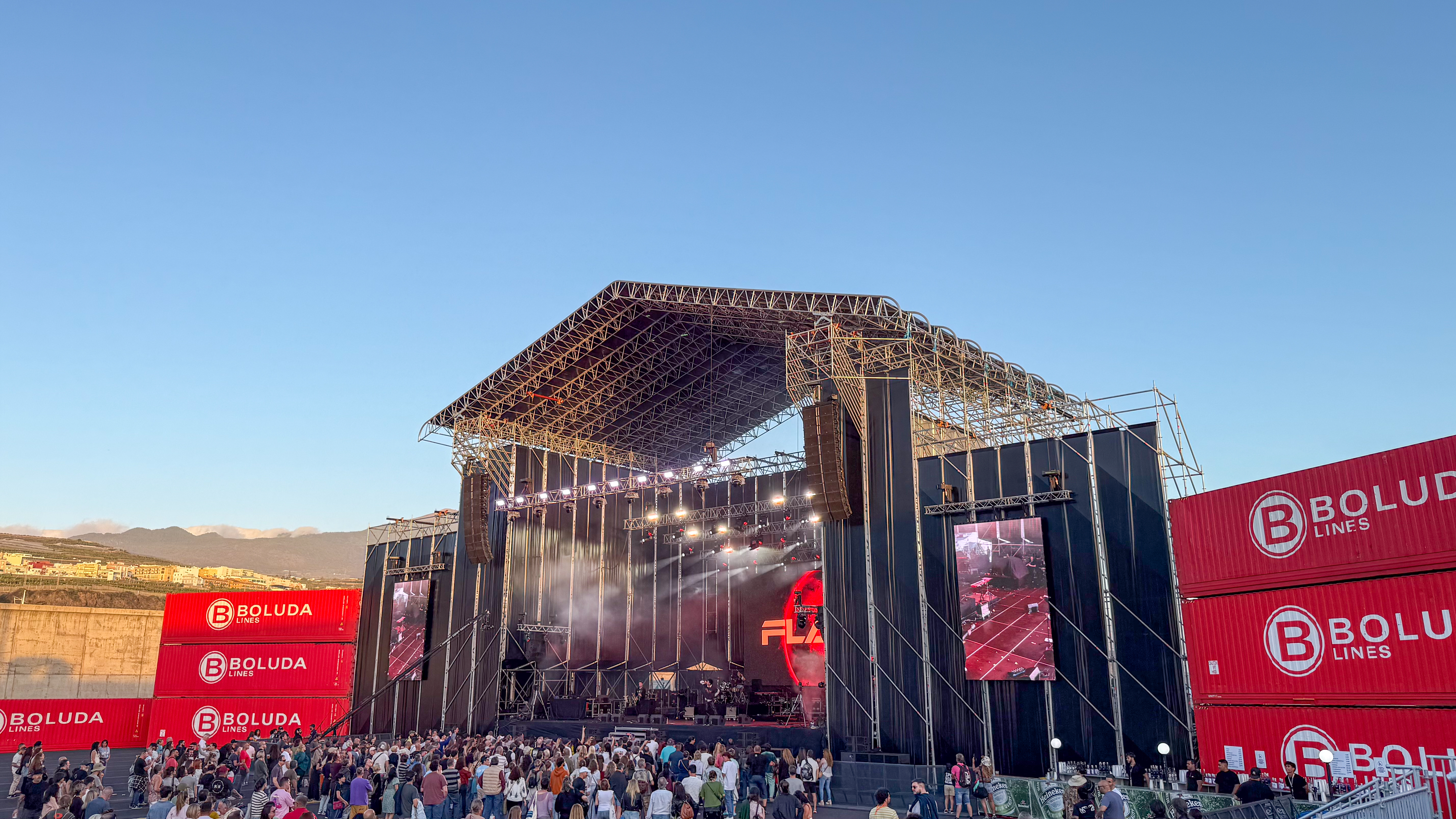 a large stage flanked either side by red shipping containers, distant mountains appear in the left of the image and a blue sky above.