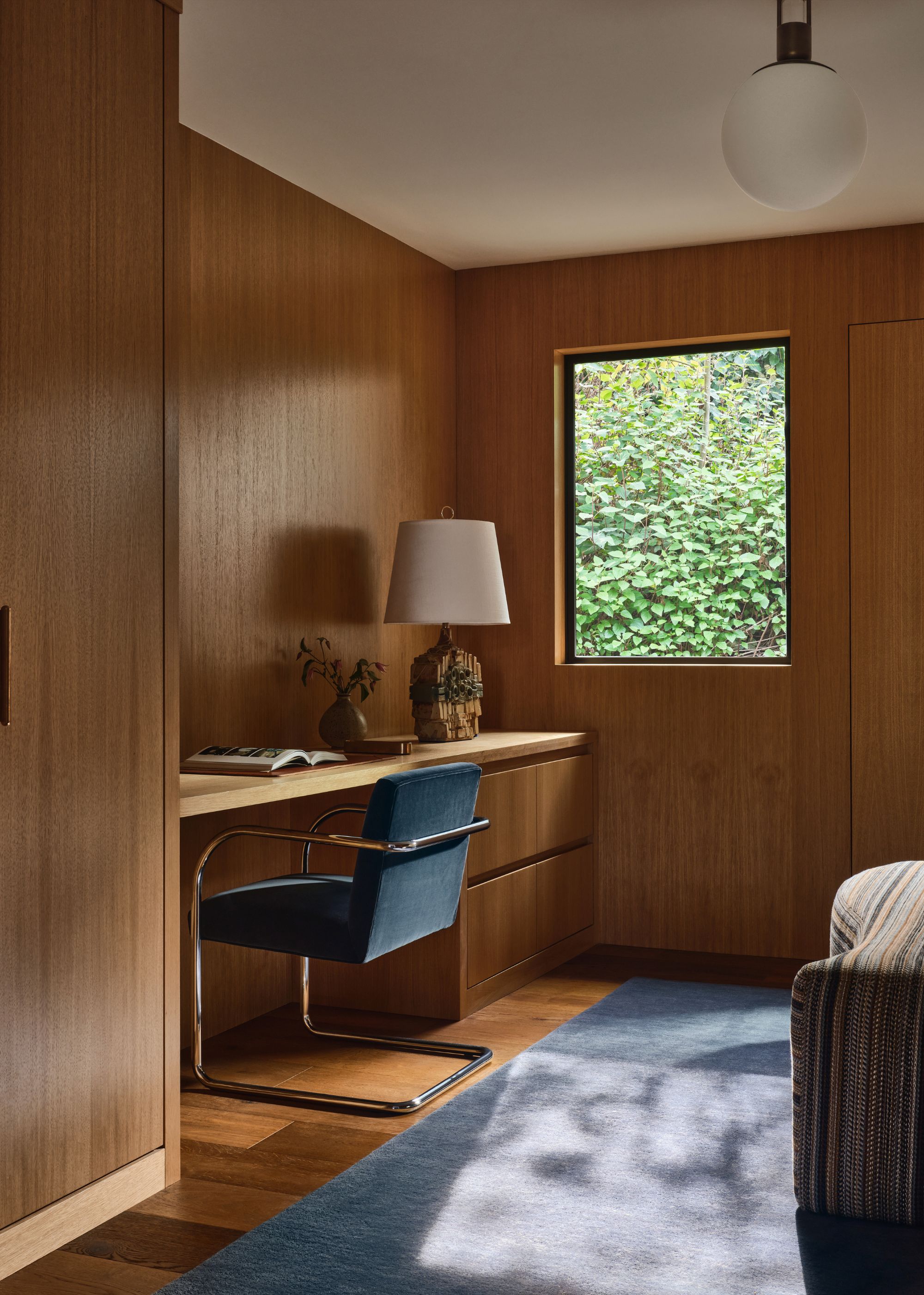 A wood-clad room with a built-in desk and a blue desk chair, a blue rug and a window.