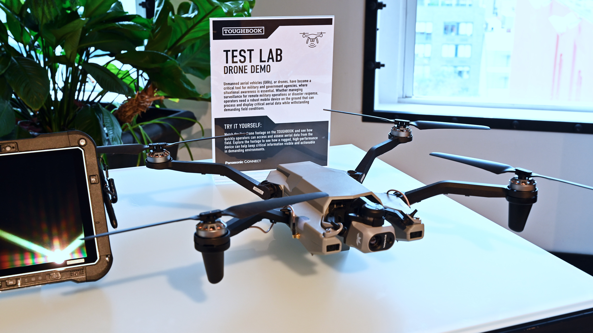 A gray quadcopter drone on a desk next to a "TEST LAB DRONE DEMO" sign and a Panasonic TOUGHBOOK screen.